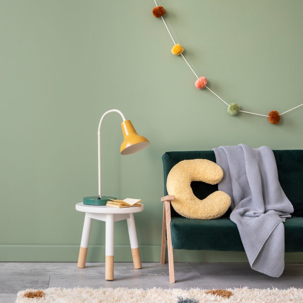 Creative composition of stylish and cozy child room interior design with green wall with bottle greeen sofa, plush pilow, bright carpet, grey plaid and stool with accessories. Copy space. Template.