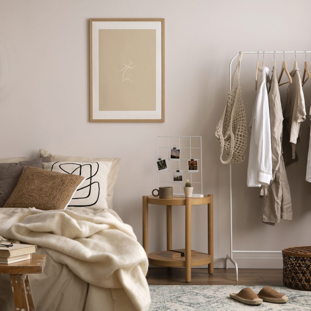 Interior design of cozy bedroom with mock up poster frame, beige bedding, stylish armchair, wooden bedside table, clothes, plants, patterned pillows and personal accessories.  Home decor. Template.